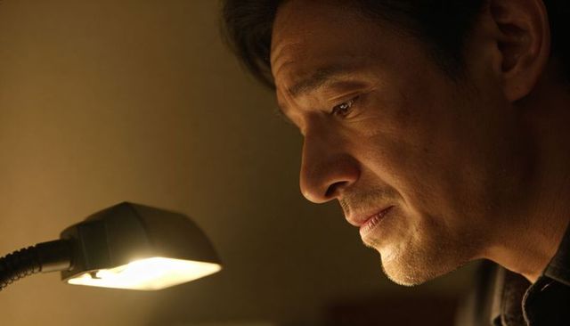 Hispanic man leaning and gazing at desk lamp, warm sidelight, pensive, wearing dark shirt