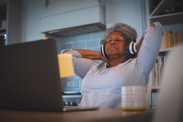 Senior African American woman sitting in kitchen, wearing headphones, and relaxing with eyes closed. Ideal for use in articles or advertisements related to senior lifestyle, relaxation, music enjoyment, home living, and mental well-being during quarantine or lockdown.