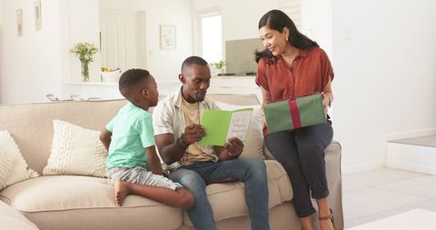Father reading father's day card while partner presenting wrapped gift with son on couch