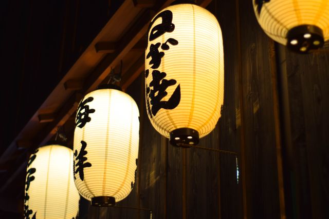 Japanese paper lanterns hanging from the ceiling, illuminating a traditional restaurant with warm light. Ideal for use in articles, blogs, travel guides about Japanese culture, traditional dining, or interior decoration inspiration. Also suitable for promotional material for Japanese cuisine restaurants and cultural events.