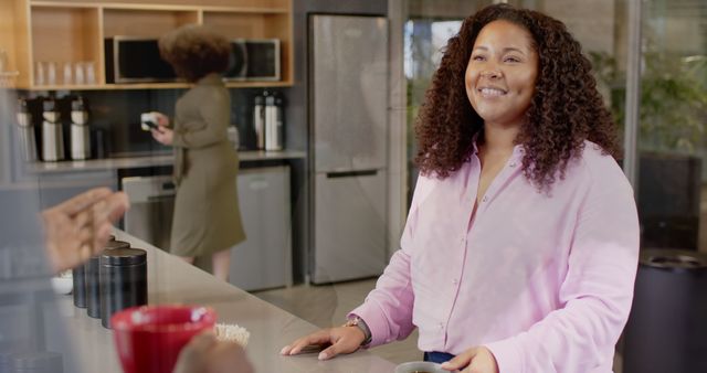 Woman is smiling while standing at a coffee counter in a modern office environment. She is wearing a pink shirt and has curly hair. In the background, another woman is preparing coffee, creating a casual and friendly atmosphere. Ideal for business, corporate environment, coworking spaces promotions, and friendly workplace culture.