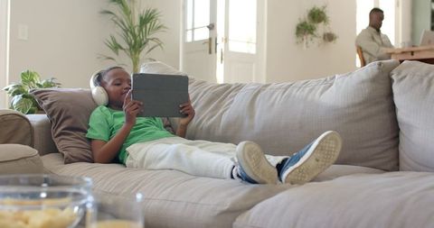 Relaxing child lying on couch holding tablet with headphones in bright modern living room