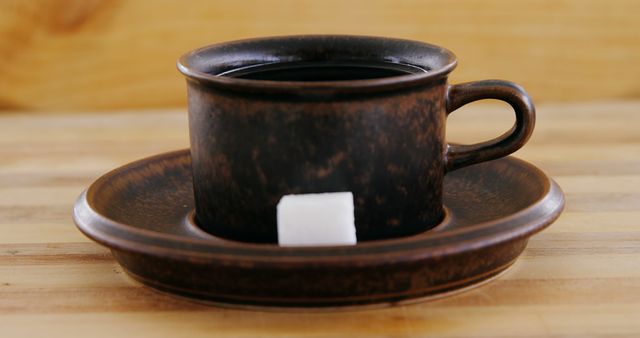 A rustic brown cup of coffee sits on a saucer with a sugar cube beside it, with copy space. Its simple presentation on a wooden table evokes a cozy, warm atmosphere ideal for a coffee break.