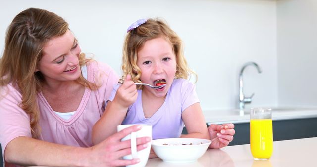 Mother feeding her young daughter breakfast at kitchen counter. Daughter is eating cereal and drinking orange juice, and both are smiling. Can be used in articles or advertisements about healthy eating, parenting, family morning routines, and fostering family bonds.