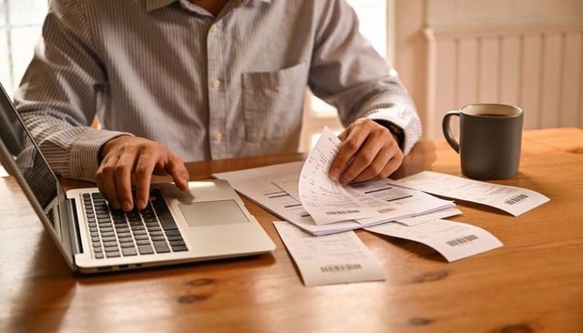 Man sorting receipts and using laptop for home accounting and expense tracking with mug