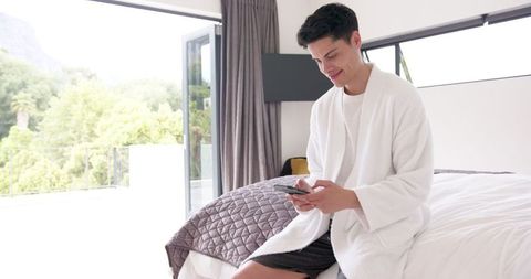 Relaxed Man Sitting on Bed Using Smartphone