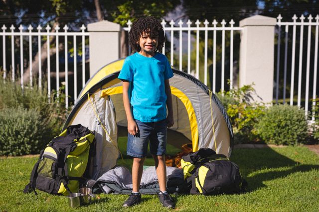 Full length portrait of smiling hispanic boy standing against tent and backpacks at backyard camp. unaltered, childhood and camping concept.
