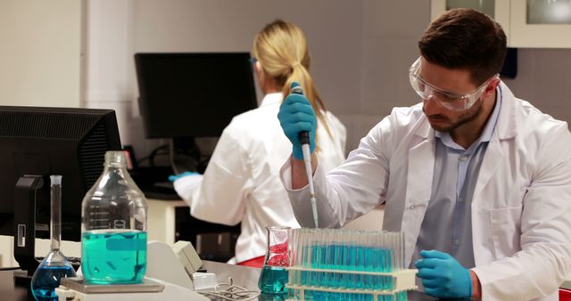 Scientists in a laboratory working with blue liquid in test tubes and various lab equipment. One scientist is using a pipette while the other works on a computer in the background. Ideal for illustrating scientific research, laboratory practices, medical experiments, and educational content on science.