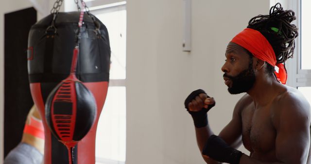 African American boxer training intensely with a punching bag. He is wearing boxing gloves and a red headband, showing strong focus and determination in the gym. Ideal for use in articles and advertisements about fitness, boxing training, sports motivation, and gym equipment promotions.