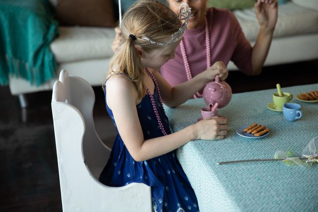 Caucasian woman spending family time together with her daughter at home, the girl wearing crown pouring tea having dolls tea party. Family leisure time at home.