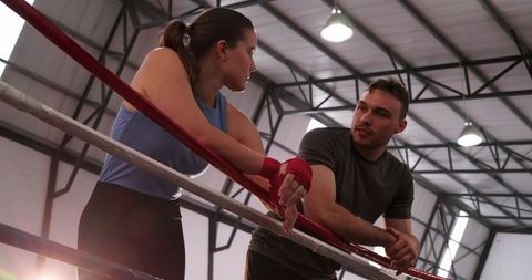 Female boxer and trainer leaning on ring ropes, resting and talking in industrial gym