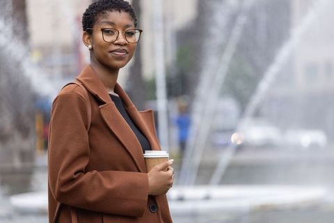 Confident african woman standing by urban fountain holding takeaway coffee, wearing brown coat