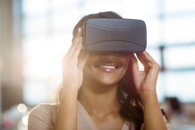 Businesswoman using virtual reality headset in office, smiling and engaging with immersive technology. Ideal for illustrating modern workplace innovation, tech-savvy professionals, and the use of VR in business settings. Suitable for articles, blogs, and marketing materials related to technology, virtual reality, and professional environments.