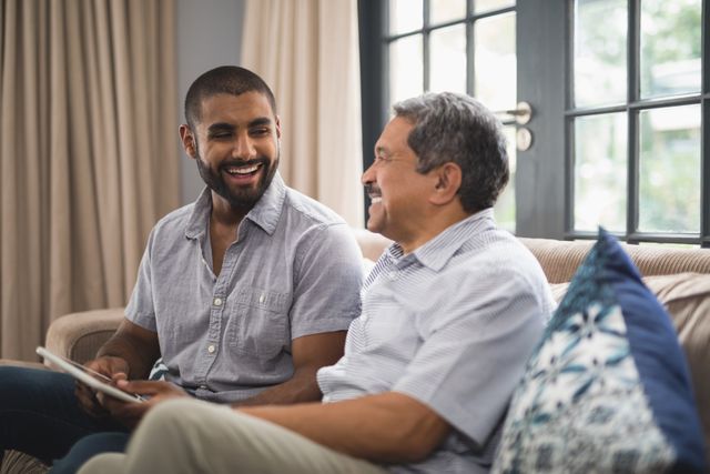 Father and son sitting together on couch at home, smiling and enjoying each other's company. Ideal for use in family-oriented advertisements, articles on family relationships, or promotional materials for home and lifestyle products.