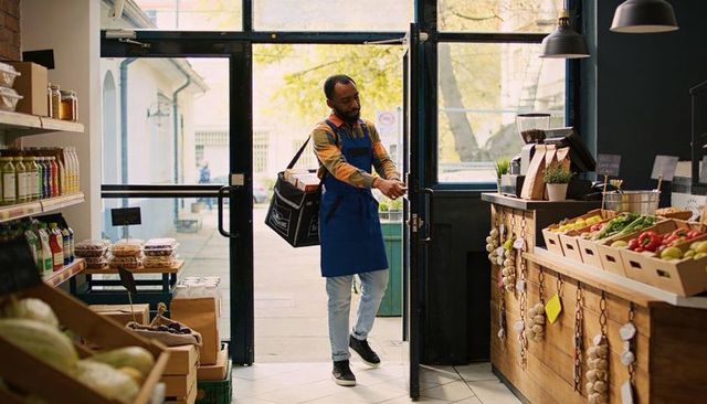 Grocery worker entering urban market carrying insulated delivery bag and fresh produce