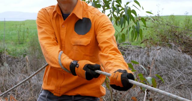 A middle-aged man in an orange high-visibility shirt is working outdoors, gripping a metal rod with gloves, with copy space. His attire suggests he might be a construction worker or involved in manual outdoor labor.