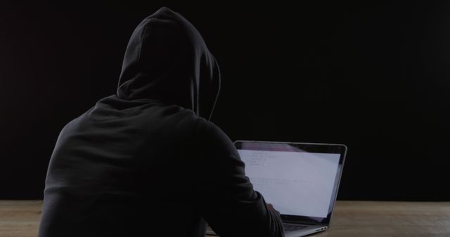 Hooded figure works on a laptop in a dimly lit room. The secretive ambiance suggests a focus on privacy or cybersecurity concerns.
