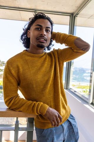 Indian man leaning on balcony rail wearing mustard sweater overlooking coastal view