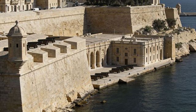 Sunlit limestone fortress overlooking harbor with row of historic cannons on quay