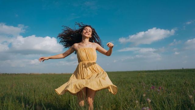 Joyful woman spinning in yellow sundress on wildflower meadow under dramatic blue sky