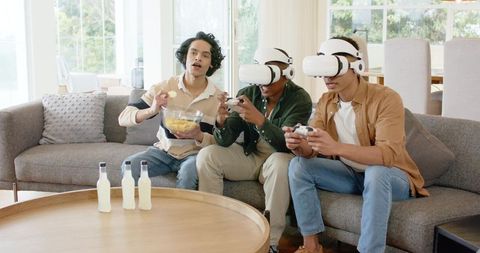 Multicultural friends playing virtual reality games on couch while snacking and laughing
