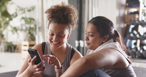 Sunlit gym friends sharing smartphone and fitness progress, relaxed diverse women duo