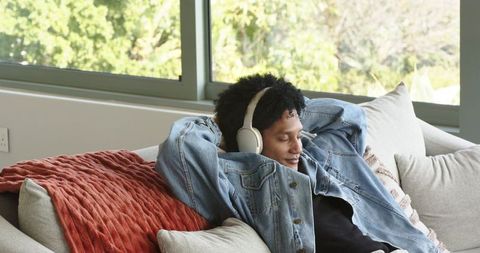 Mid-adult african american man relaxing on sunlit sofa listening to music with headphones