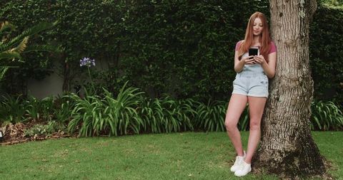 Redhead woman leaning on tree texting on smartphone in lush backyard garden