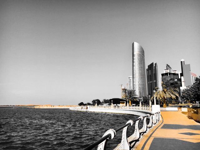 This scenic view highlights the sleek, modern skyline of Abu Dhabi with the clear waters and a sunny walkway. Ideal for use in travel blogs, architectural features, or promotional materials showcasing urban development and tourism in the Middle East.
