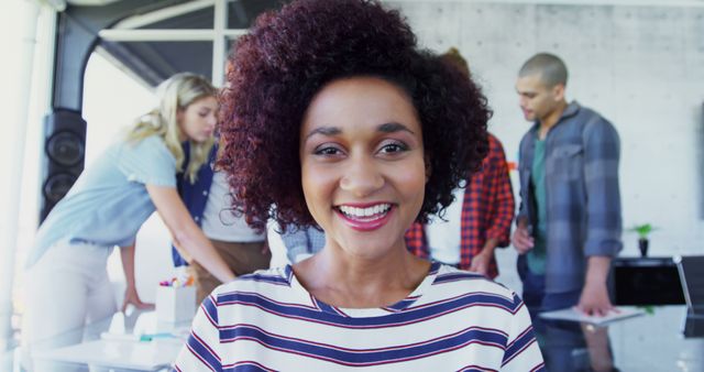 Young woman with curly hair smiling in a modern office environment. In the background, her colleagues are working together and collaborating on a project. This image is excellent for showcasing diversity, modern workplace culture, ideas of teamwork, or advertising on professional networking websites.