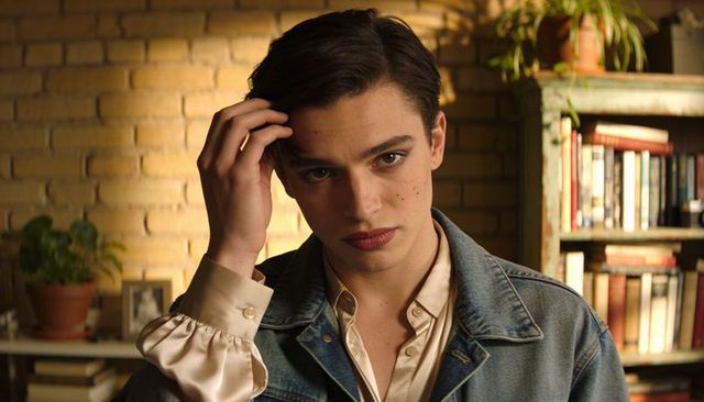 Intimate portrait of man touching hair in denim jacket and satin shirt, warm bookcase glow