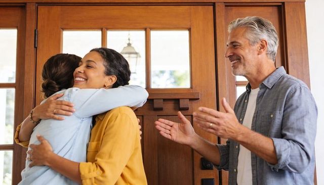 Women hugging at wooden front door while man welcoming and smiling sunlit warm home reunion