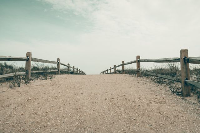 Image showcases a serene coastal path leading to the beach, flanked by a wooden fence. Clear sky adds to the peaceful ambiance. Useful for travel promotion, relaxation themes, nature blogs, and vacation advertisements.