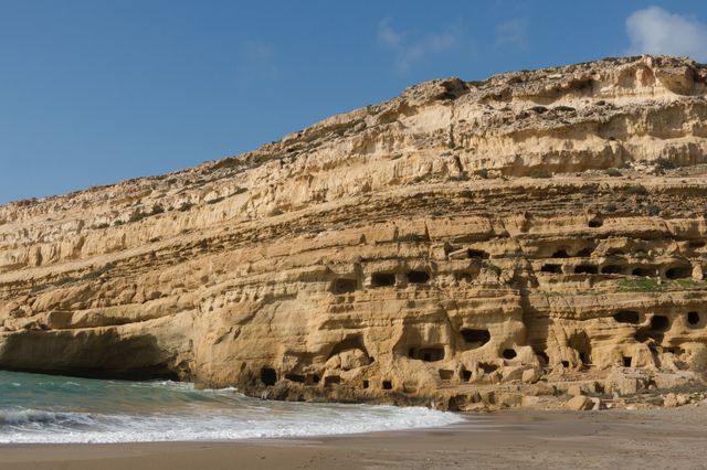 This image captures Matala Beach on the island of Crete, Greece, featuring sandstone cliffs with numerous caves. The azure sea waves gently against the sandy shore, framed by a clear blue sky. Ideal for travel brochures, magazines about historic sites, nature blogs, and materials promoting Greek tourism.