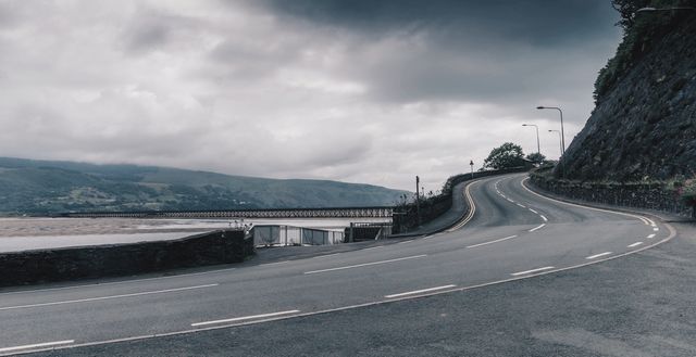 Scene depicting an empty winding coastal road curving alongside a rocky cliff under an overcast sky. Ideal for backgrounds, travel magazines, road trip promotions, blog illustrations, and atmospheric settings conveying solitude and anticipation.