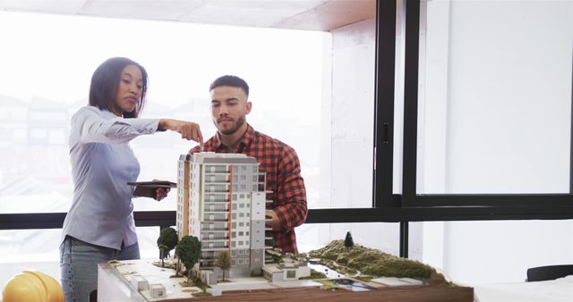 Architects discussing and pointing at details of a building model in a modern office. Ideal for content related to architectural design, collaboration, construction projects, and urban development. Can be used in articles, blogs, or websites focusing on architecture, teamwork, and professional work environments.