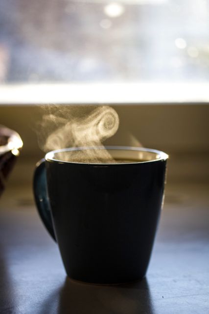Steaming mug of hot coffee placed on a window sill with soft morning light shining through. Ideal for use in articles or advertisements related to morning routines, home comfort, relaxation, and cozy atmospheres. This captures the essence of a peaceful start to the day.