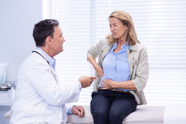 Middle-aged woman consulting with a male doctor about stomach pain in a clinical setting. The doctor is attentively listening and pointing to the area of discomfort. This image can be used for healthcare, medical advice, patient care, and health-related articles or advertisements.