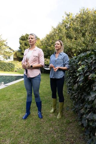 Two middle-aged women standing poolside wearing knee-high boots holding garden shears