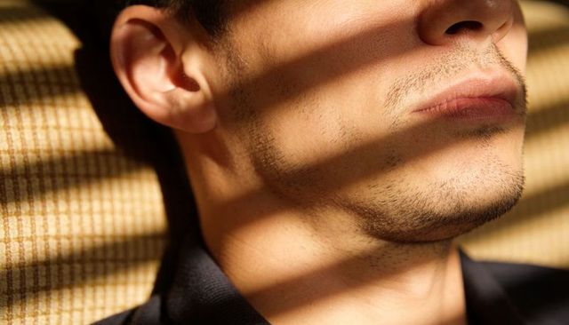 Male jawline showing light stubble and window blind shadow stripes on sofa closeup