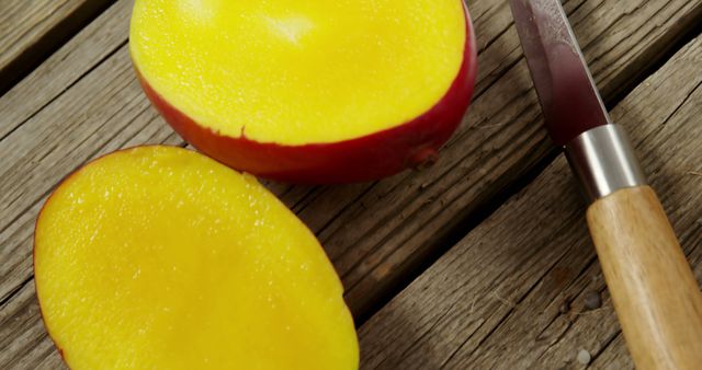 Close-up showing two halves of ripe, juicy mango on weathered wood table, with knife nearby. Captures vibrant yellow color and appetizing texture of mango. Ideal for food blogs, healthy lifestyle promotions, or culinary recipe illustrations.