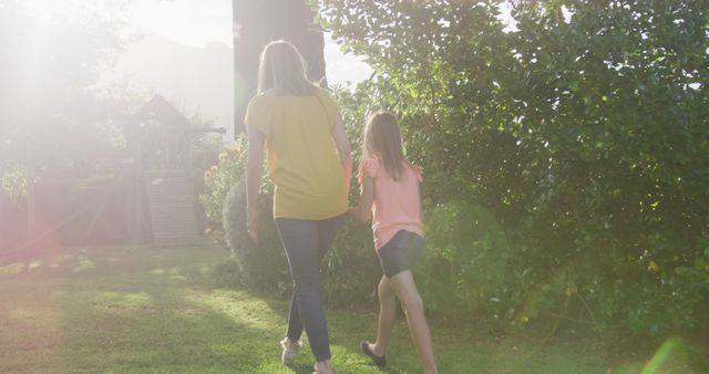 Rear view of caucasian mother and daughter walking holding hands in sunny garden, copy space. Summer, motherhood, childhood, togetherness, nature and adventure, unaltered.