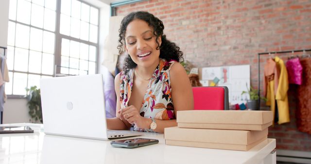 This image shows a young female entrepreneur in a modern office environment working on her laptop. She is smiling and appears to be happy and productive. There are package boxes and a phone on the desk, indicating involvement in an online business or e-commerce. Great for topics related to entrepreneurship, small business, modern office setups, and business technology use.