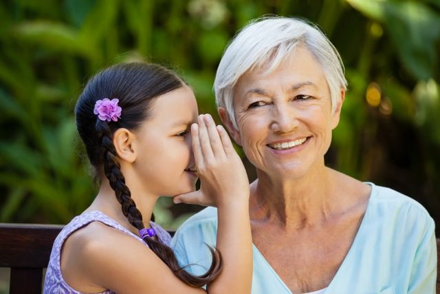 Granddaughter whispering to smiling grandmother in backyard, showcasing family bonding and love. Ideal for use in advertisements, family-oriented content, and articles about intergenerational relationships, happiness, and outdoor activities.