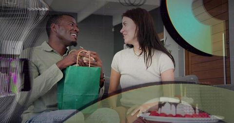 Black man presenting green gift bag to white woman on couch with lit birthday cake