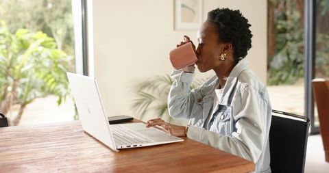 African American Woman Working Remotely on Laptop Drinking Coffee in Bright Home Office