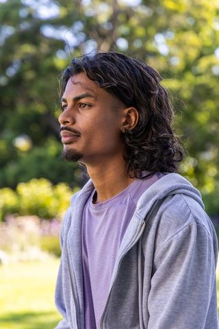 Contemplative young man standing in sunlit park wearing grey hoodie and hoop earring