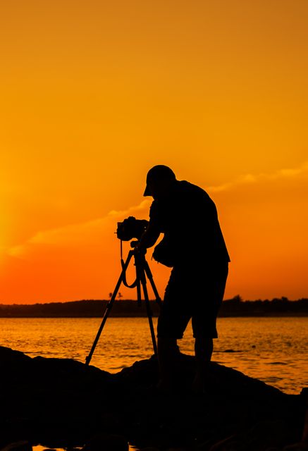 Silhouette of male photographer adjusting camera on tripod while standing on rocky beach during sunset. Beautiful golden sky casts a warm glow over the scene. Ideal for concepts of professional photography, hobbies, artistic pursuits, and capturing nature's beauty. Also suitable for travel, leisure, and lifestyle themes.