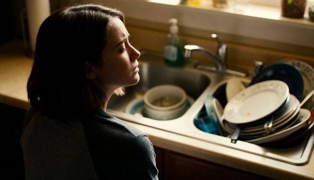 Young woman pausing at sink with stacked dishes in sunlit kitchen, contemplative mood