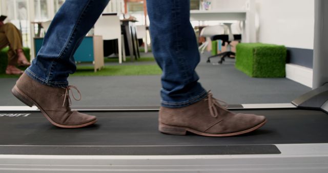 This image depicts a person walking on a treadmill in an office environment. The individual is wearing casual shoes and jeans, indicating a relaxed yet professional setting. Green, grass-like carpet suggests an eco-friendly or outdoor-themed workspace. This image can be used to illustrate concepts such as workplace wellness, fitness at work, modern office designs, promoting a healthy lifestyle among employees, or innovative business environments.